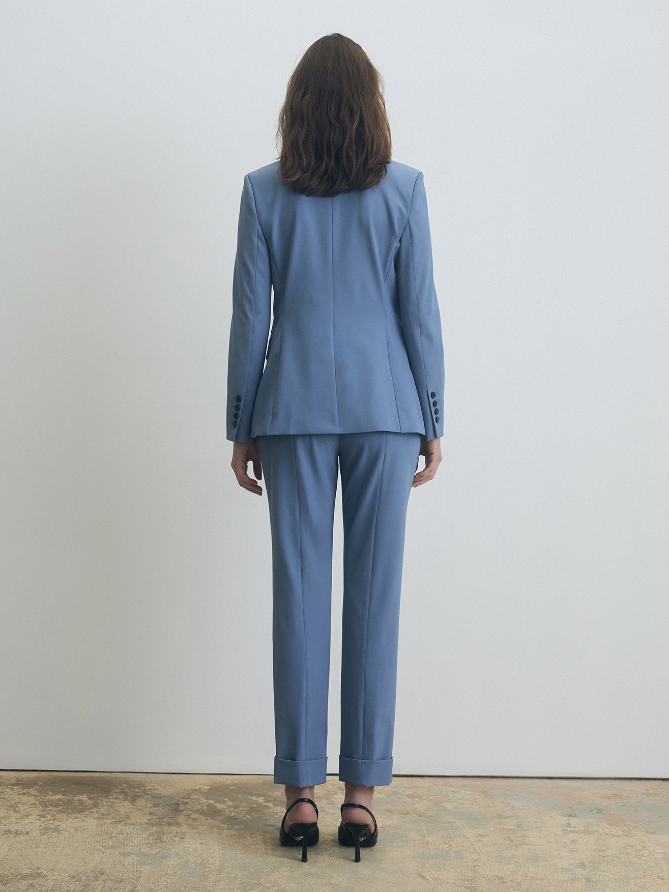 Rear view of a smokey blue tailored suit showing the structured back of the single-breasted jacket with buttoned cuffs, paired with straight-leg trousers with turned-up hems and black stiletto heels.