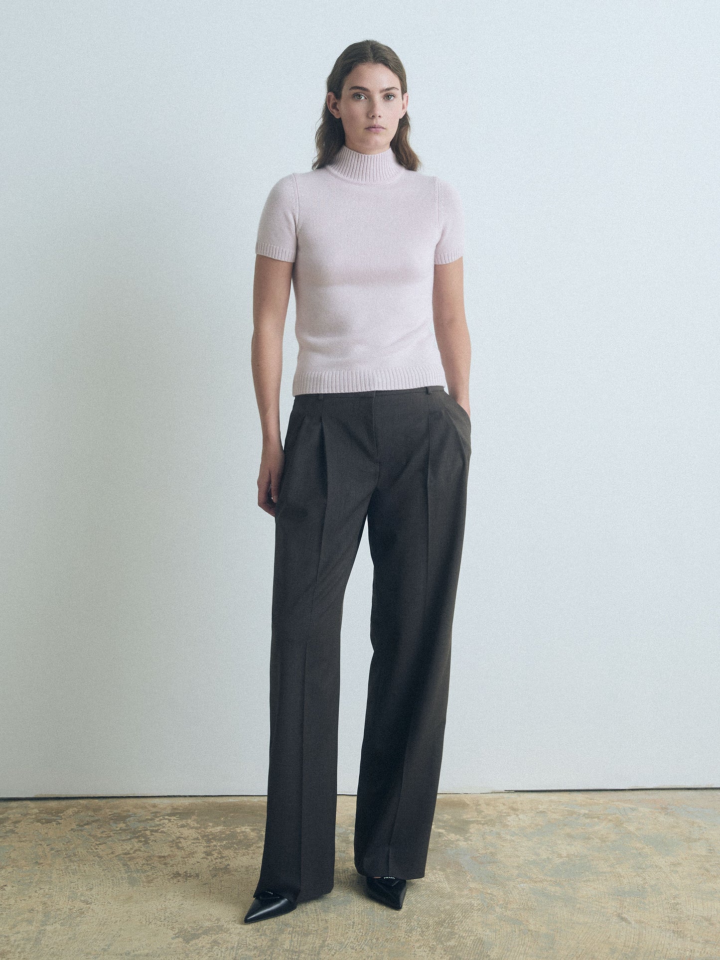 Woman wearing a light pink judith and charles short sleeve turtleneck and dark gray wide-leg pants against a plain background