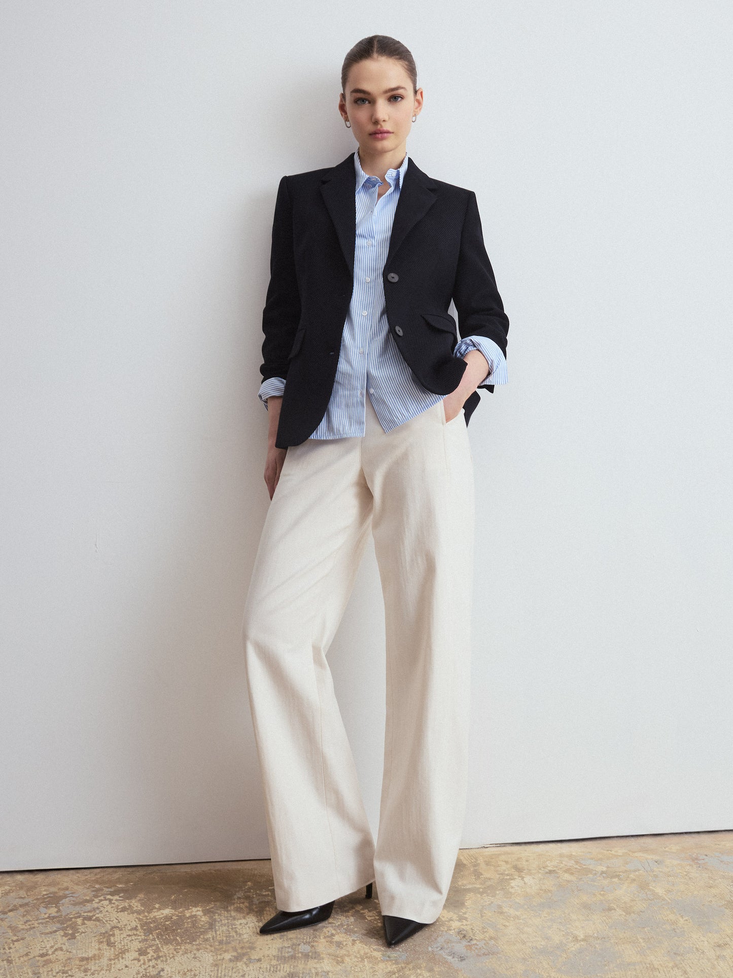 Woman wearing a navy blazer, light striped blue shirt, and white pants against a plain background