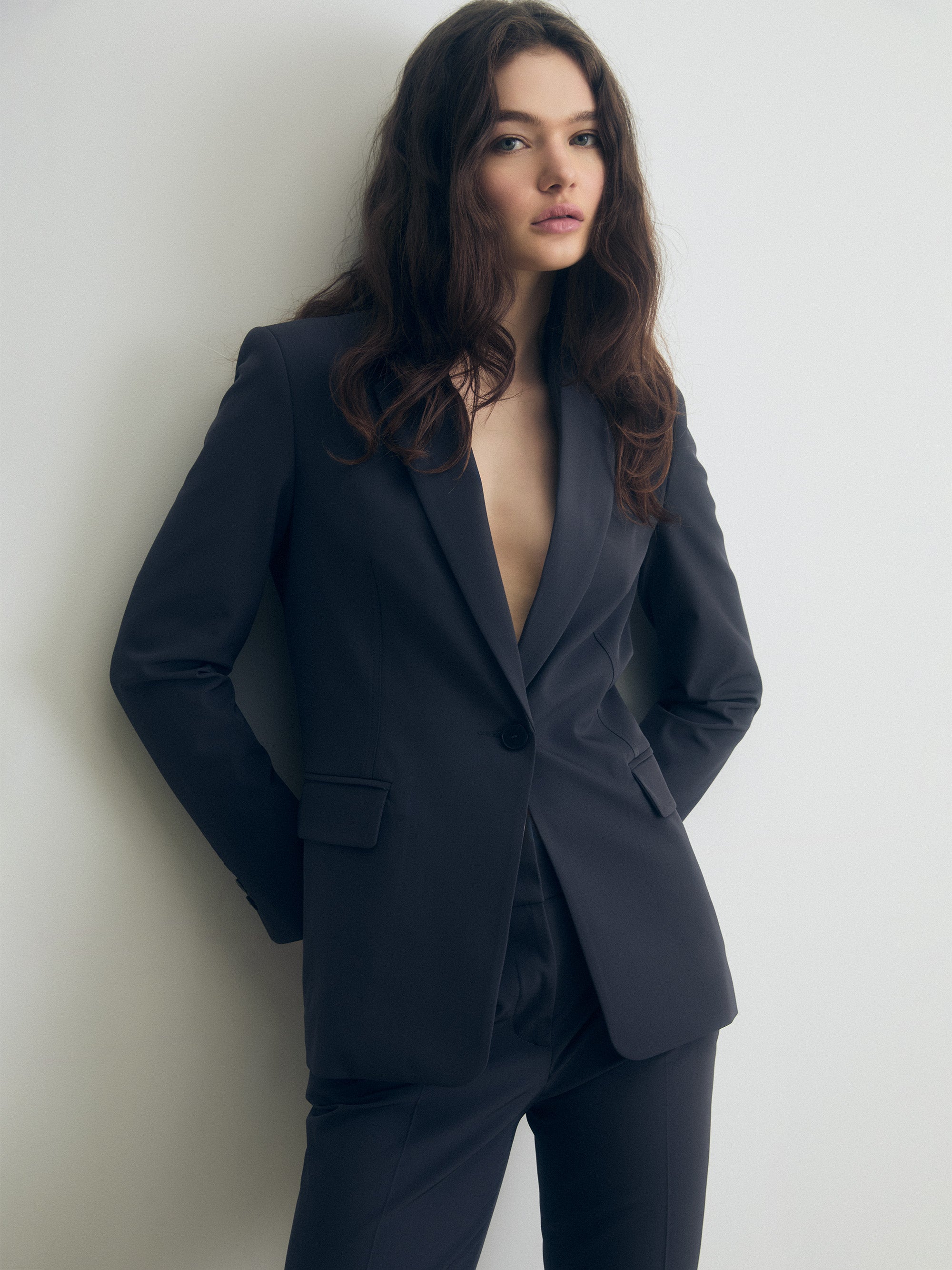 Tailored dark grey jacket with a single-breasted design, sharp peak lapels, and structured shoulders, paired with matching trousers. The jacket features a single button closure and flap pockets, creating a sleek, modern silhouette.