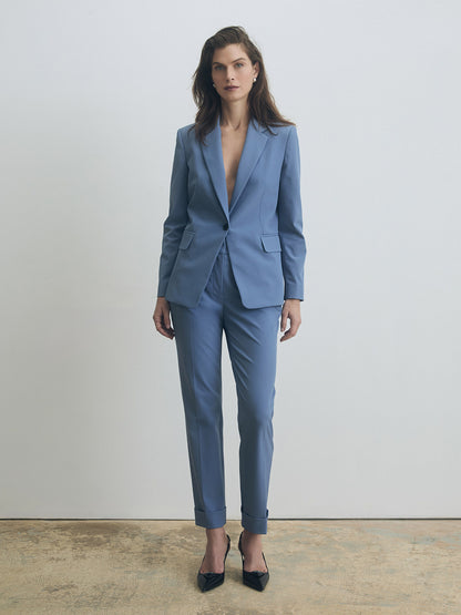 Full-length view of a tailored smokey blue suit featuring a single-breasted jacket with peak lapels, flap pockets, and a single button closure, paired with slim, cuffed trousers and black pointed-toe heels.