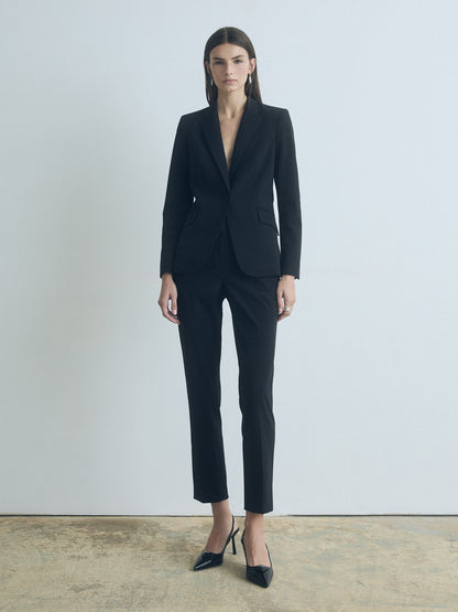 A model wearing black ankle length slim black pants with mid-rise waist, featuring slash pockets, back welt pockets, hook and eye closure with zip, and belt loops.