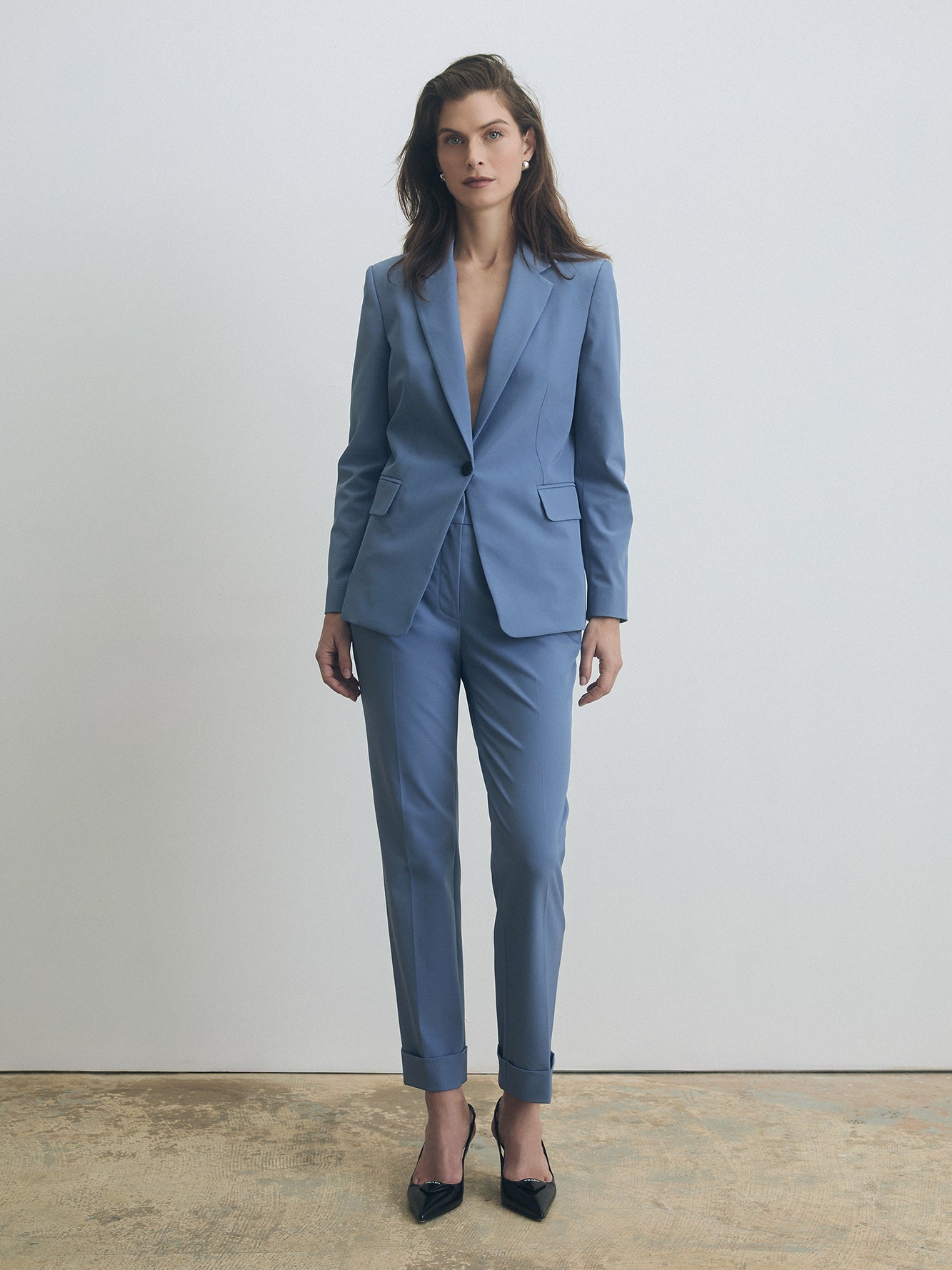 Model wearing a tailored smokey‑blue pantsuit featuring a fitted single‑button blazer with notched lapels and flap pockets, paired with slim cuffed trousers and black pointed‑toe heels. The look is photographed against a minimal light grey background