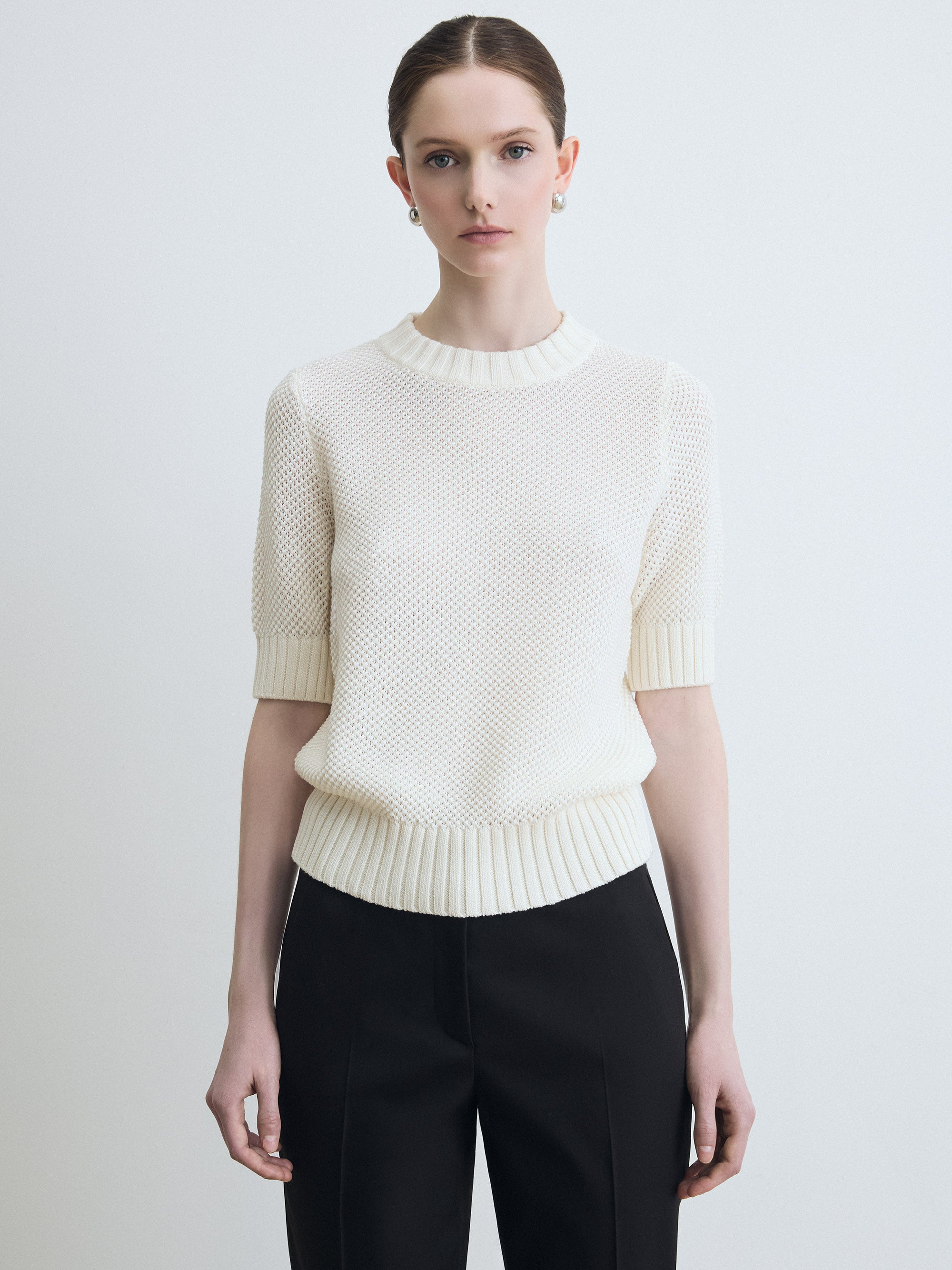 Woman wearing a white textured sweater and black pants on a light gray background