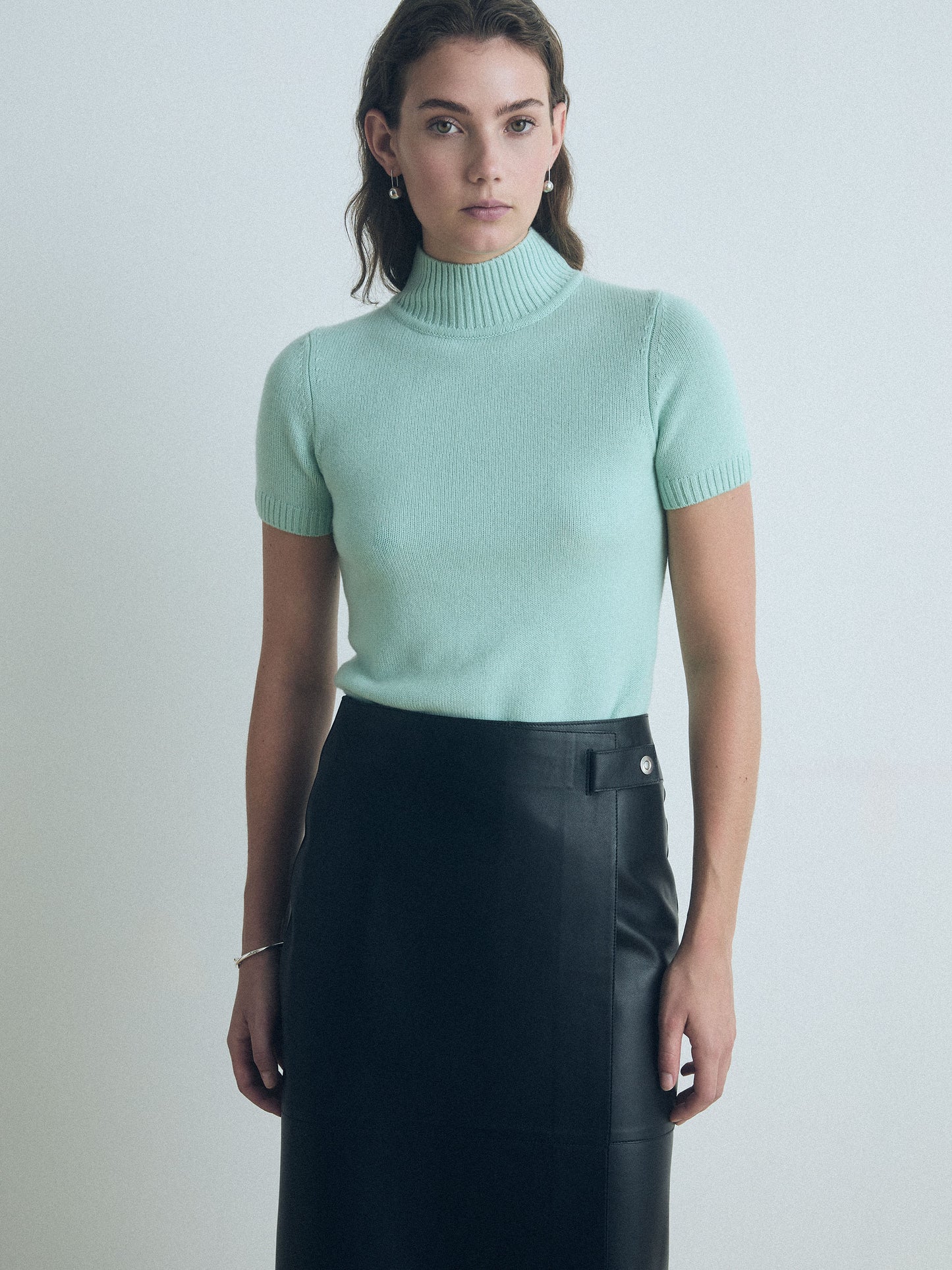 Woman wearing a light green short sleeve judith and charles turtleneck sweater and black leather skirt against a plain background