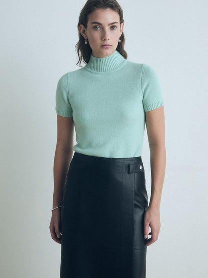 Woman wearing a light green short sleeve judith and charles turtleneck sweater and black leather skirt against a plain background