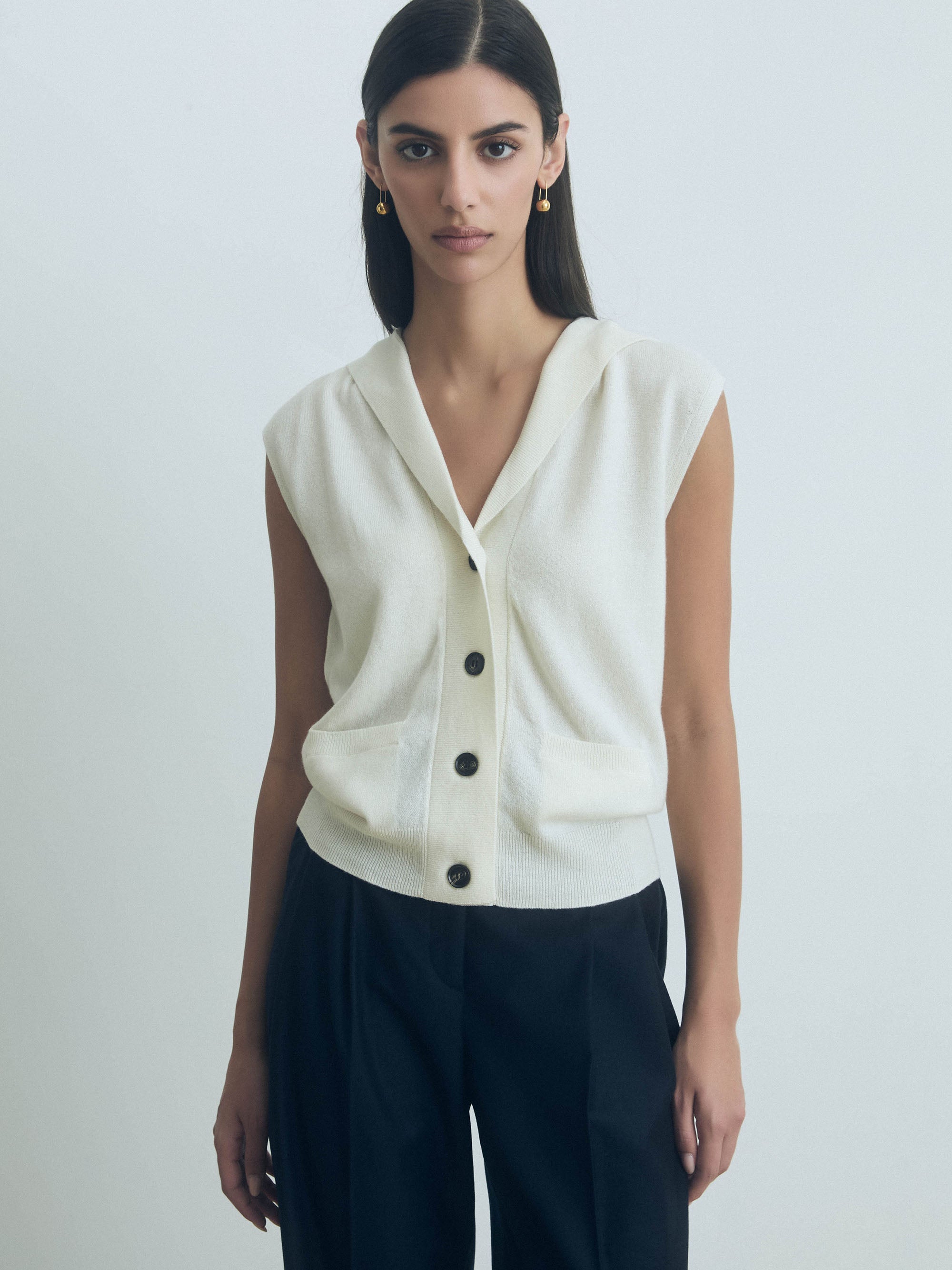 Woman wearing a Off White sleeveless hooded wool-cashmere vest with button front closure by Judith & Charles with black pants on a plain background