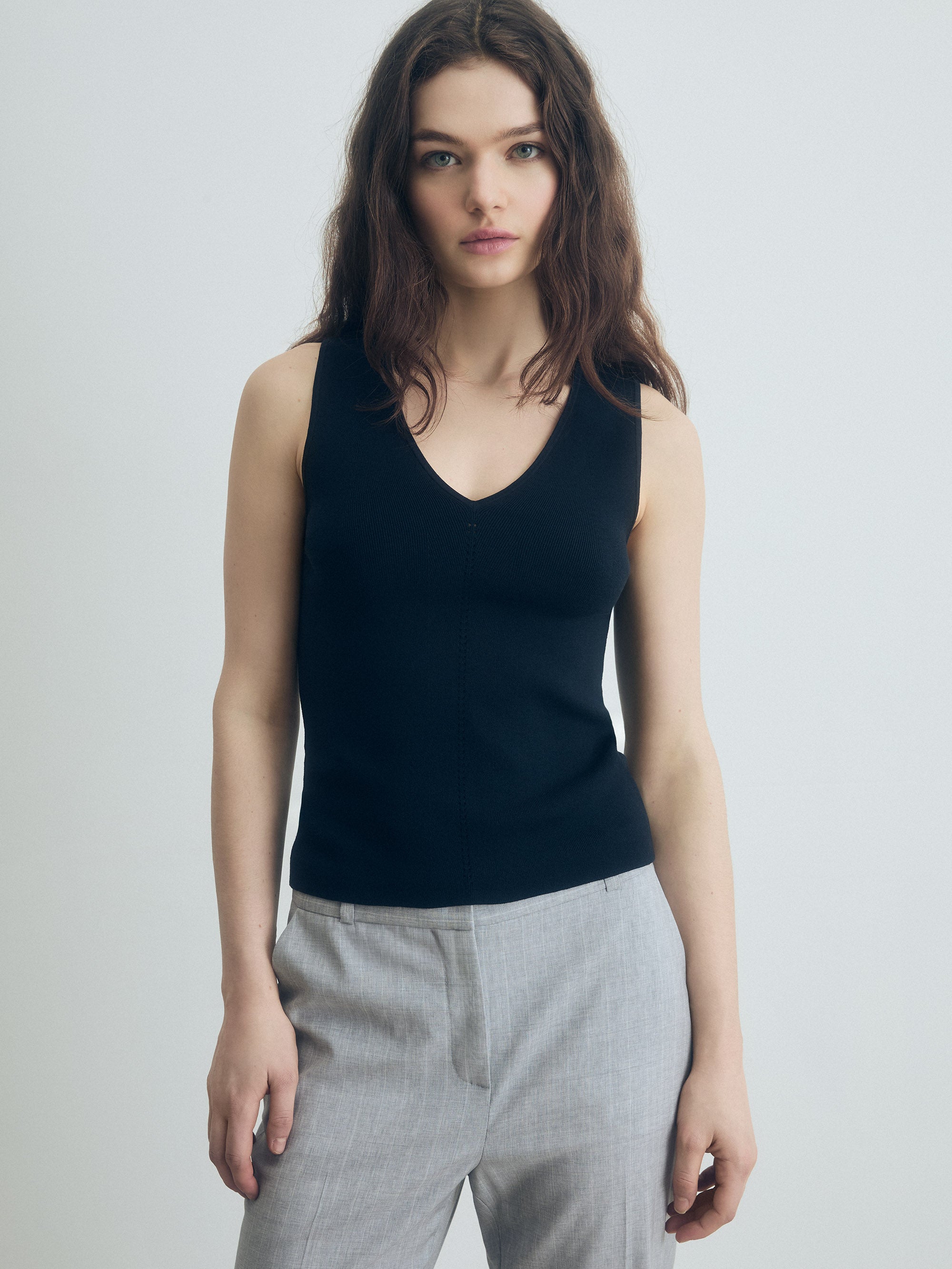 Person wearing grey trousers over a black V‑neck top, standing against a plain light background.