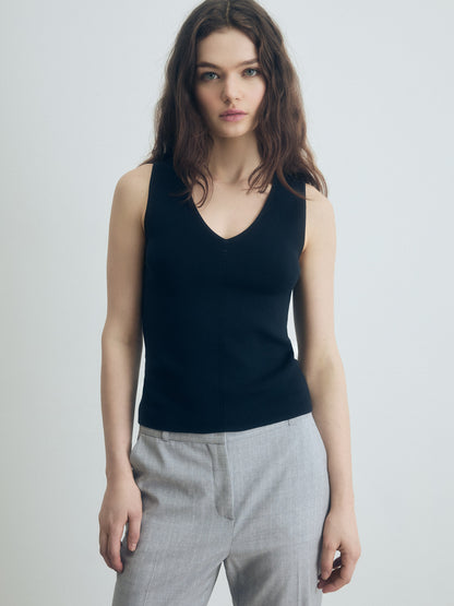 Person wearing grey trousers over a black V‑neck top, standing against a plain light background.