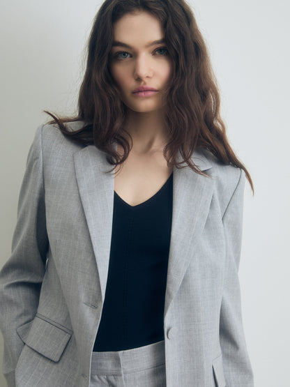 Person wearing a light grey tailored blazer and matching trousers over a black V‑neck top, standing against a plain light background.