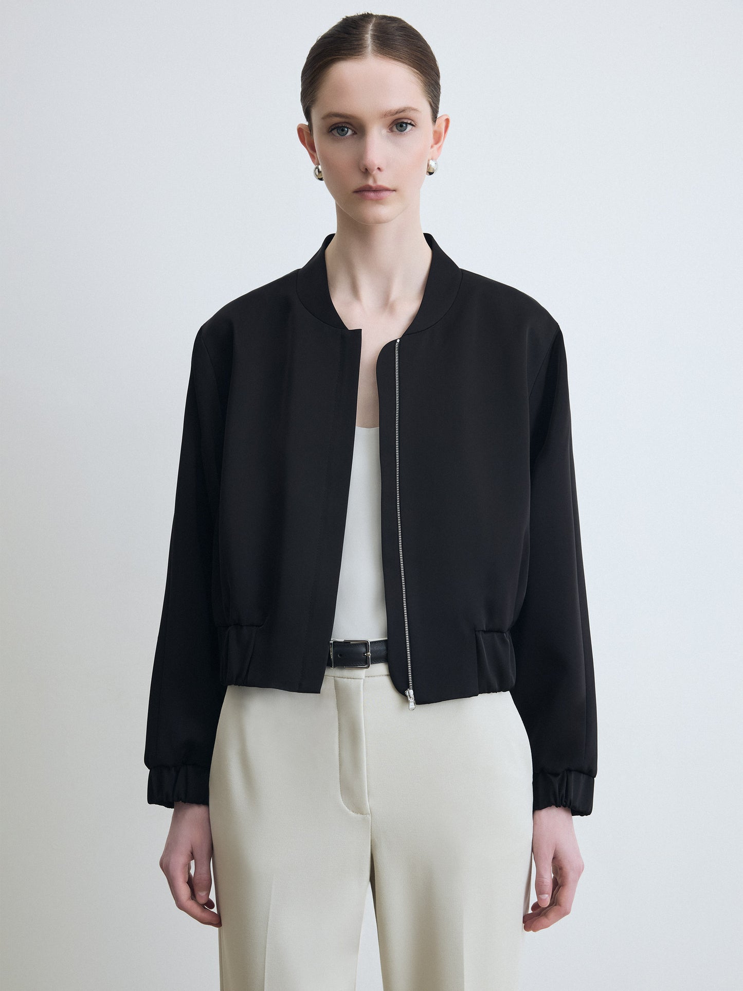 Person wearing a black bomber jacket and white pants on a light background