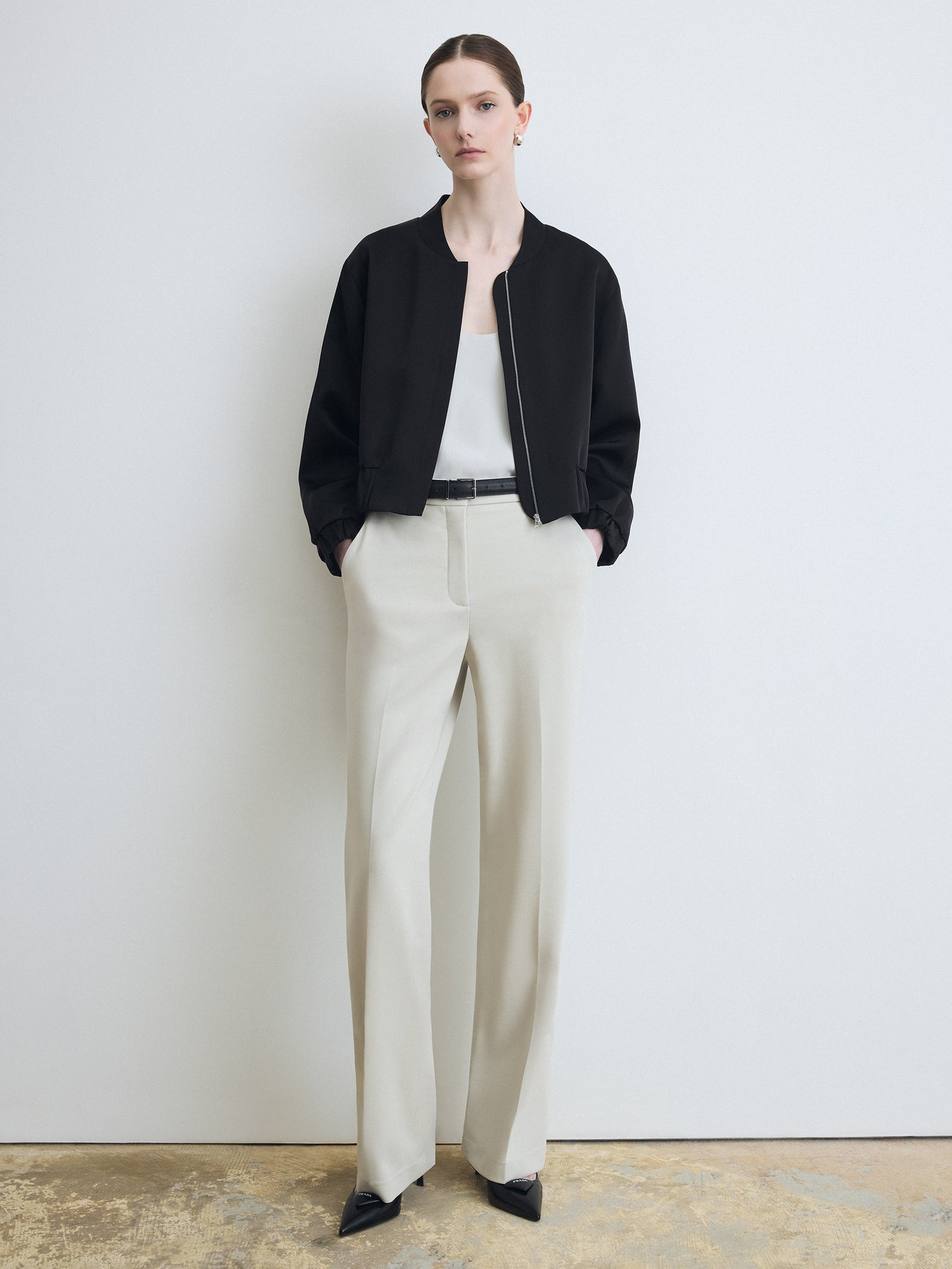 Person wearing a black jacket, white shirt, and beige pants against a plain background