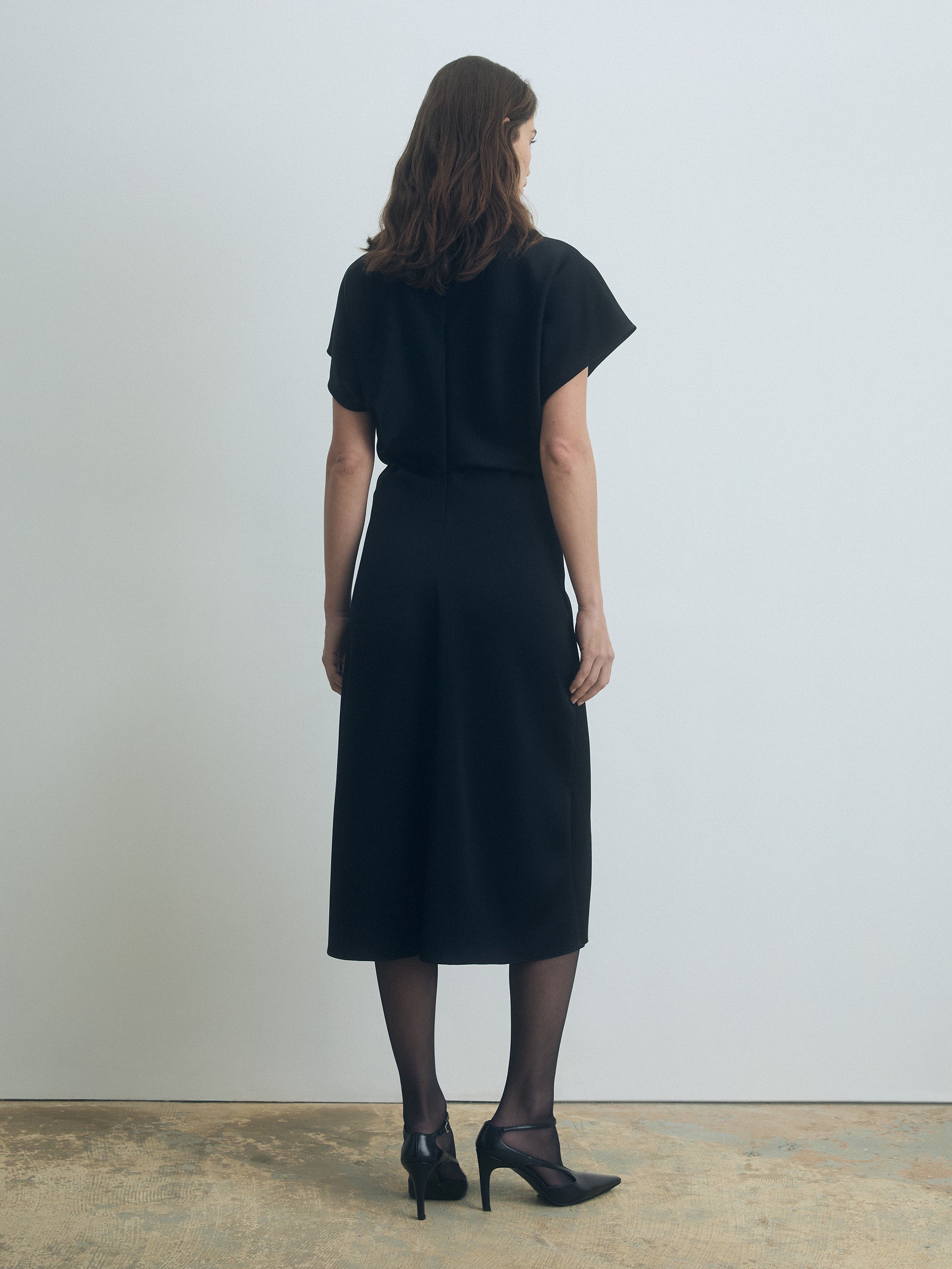 Woman wearing a black dress standing against a plain white wall.