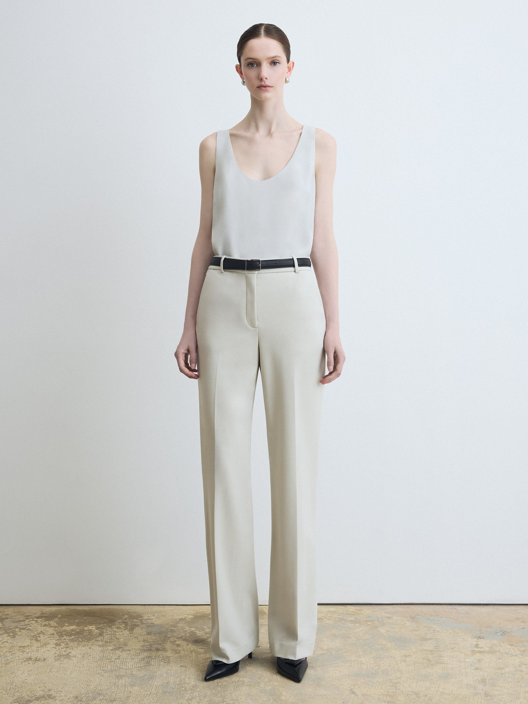 Woman wearing a white sleeveless top and beige pants against a plain background