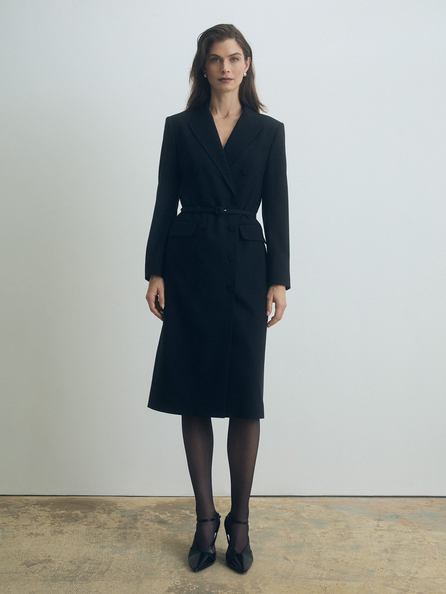 Woman wearing a long sleeved black belted double breasted coat dress standing against a plain background