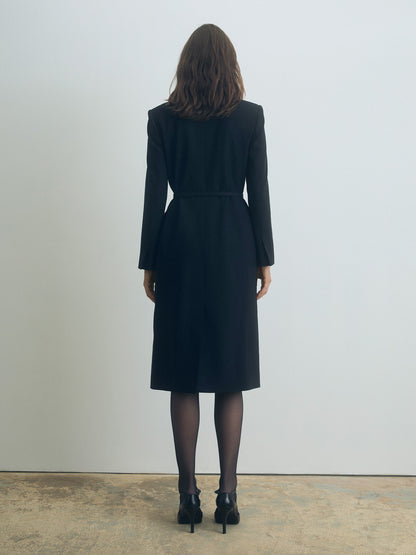 Woman wearing a Black belted double breasted dress coat standing against a plain white wall.