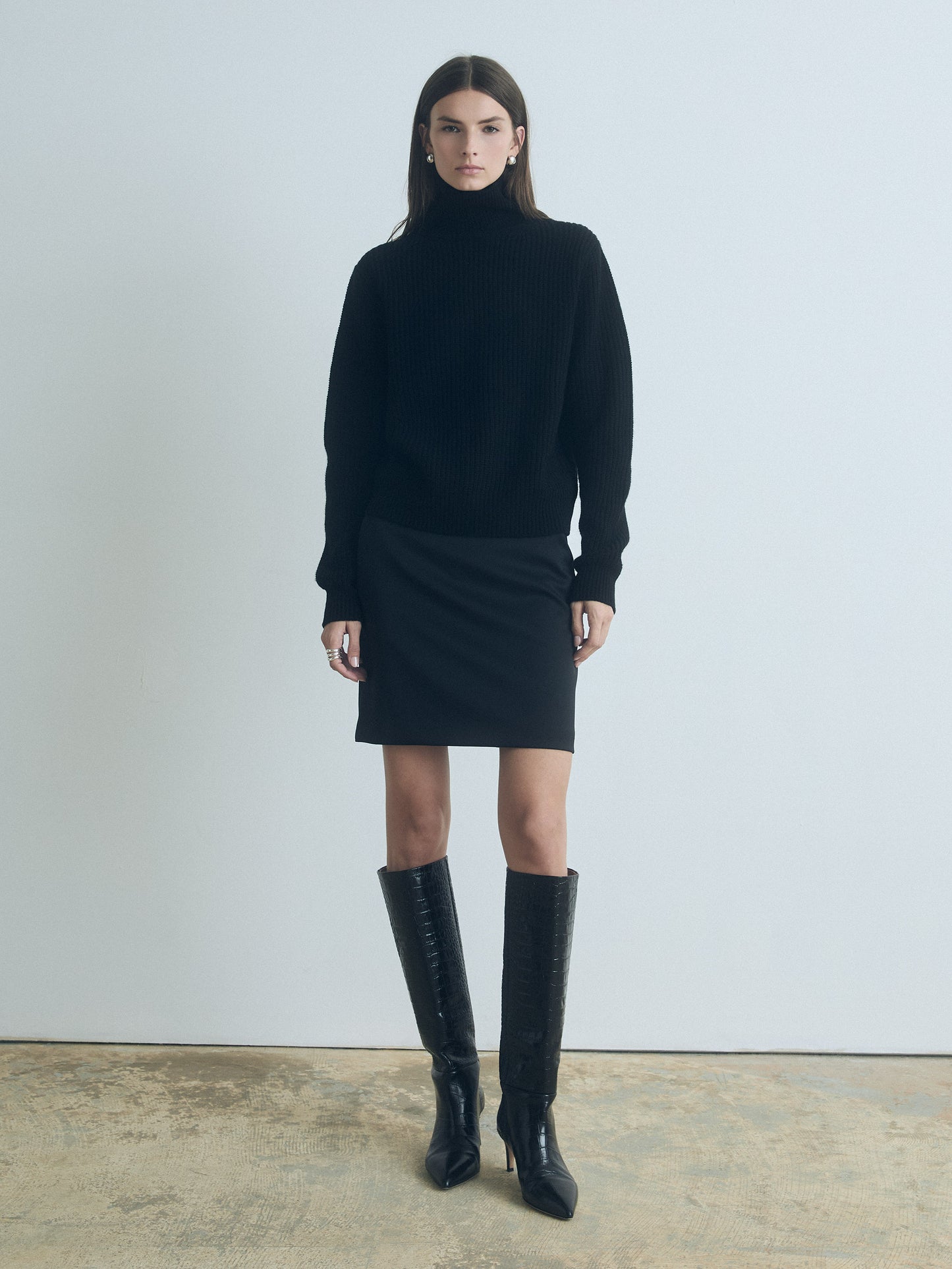 Woman wearing a black turtleneck sweater and skirt with knee-high boots against a plain background