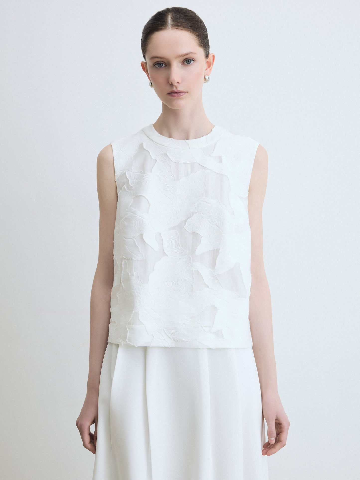 Woman wearing a floral white sleeveless top with floral details on a plain background