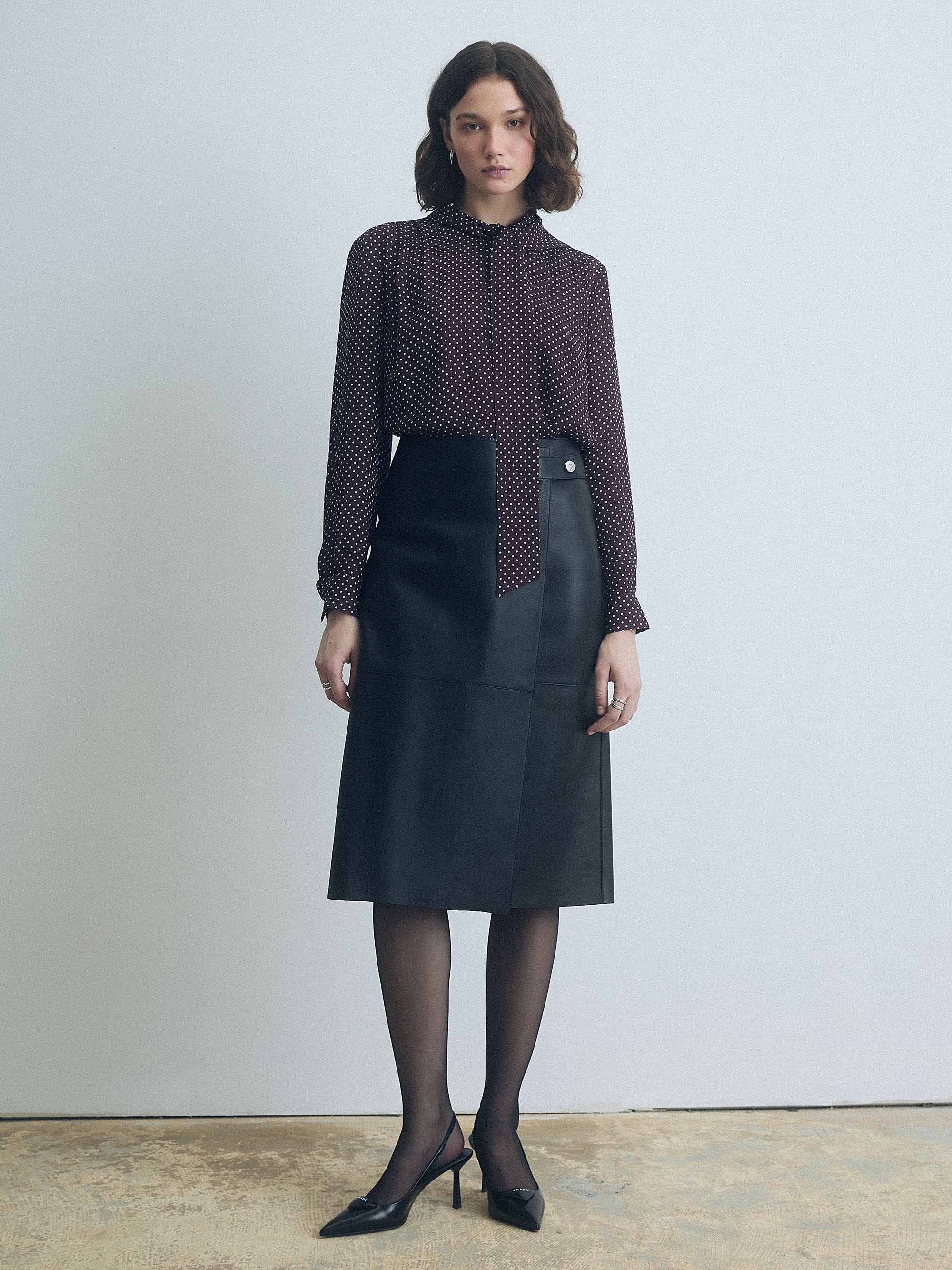 Woman wearing a dark cherry polka dot blouse with a tie by Judith and Charles  and black skirt against a plain background