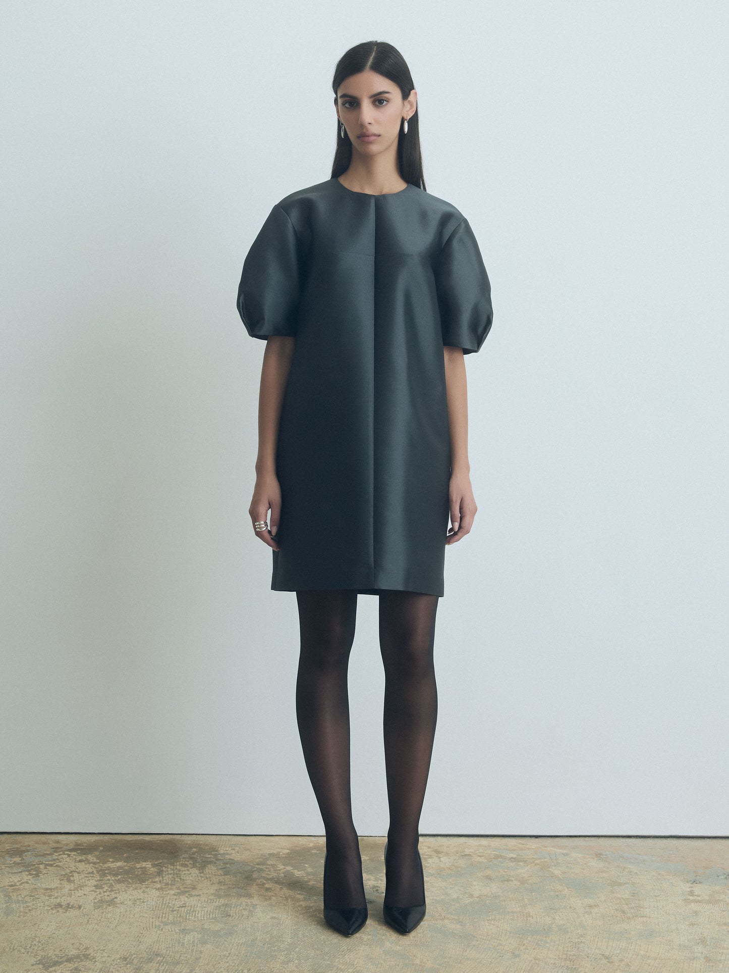Model in a crew neck dress in anthracite with short, structured sleeves and invisible back zipper against a minimalist background