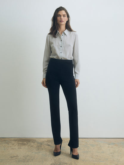 Model wearing a fitted long-sleeve blouse in black-and-white vertical stripes with a spread collar and front button closure, paired with high-waisted black tailored trousers and black pointed-toe heels
