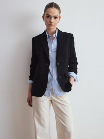 Person wearing a navy blazer over a light blue shirt with white pants against a plain background