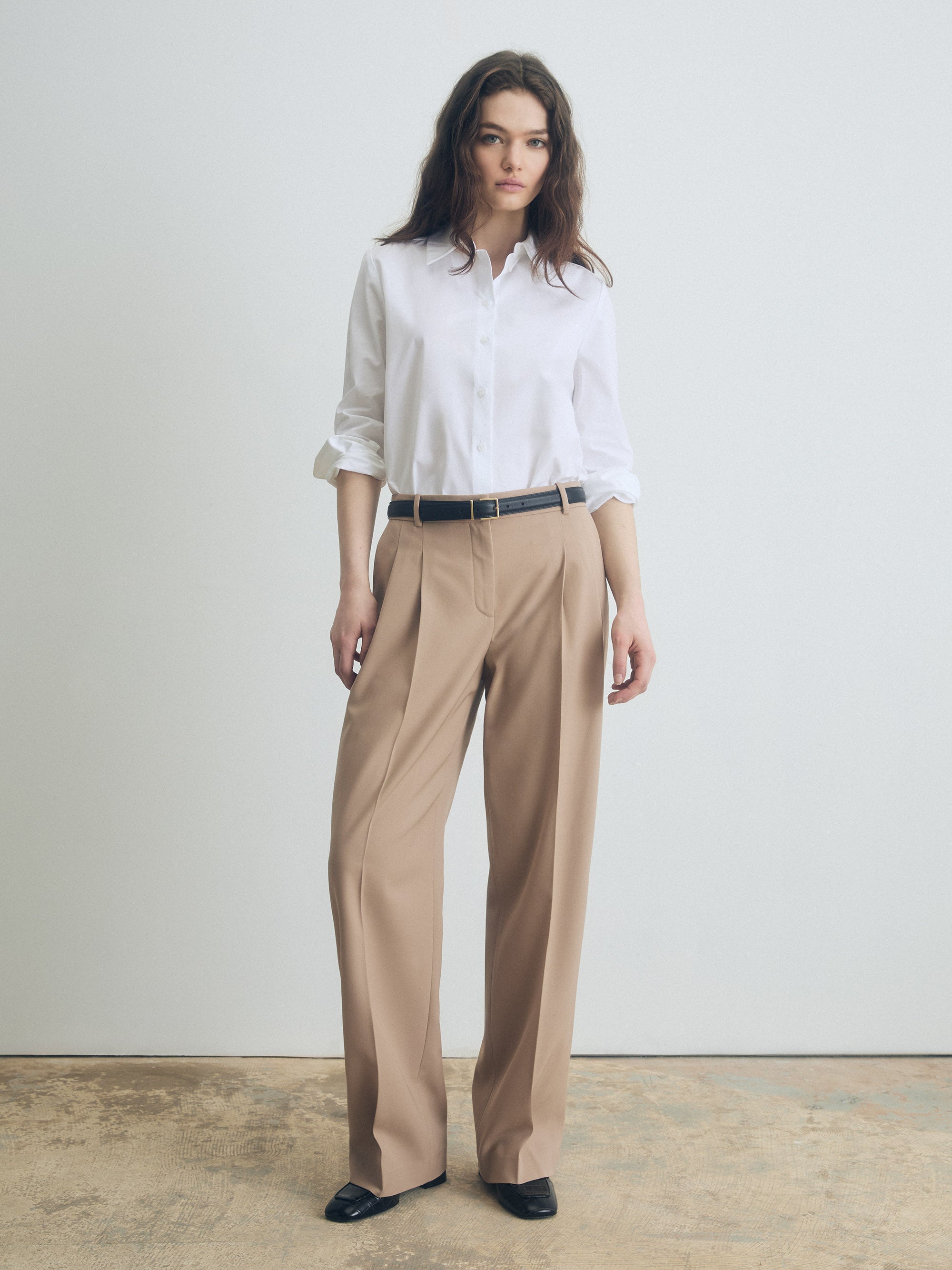 Woman wearing a white shirt and camel pants against a plain background