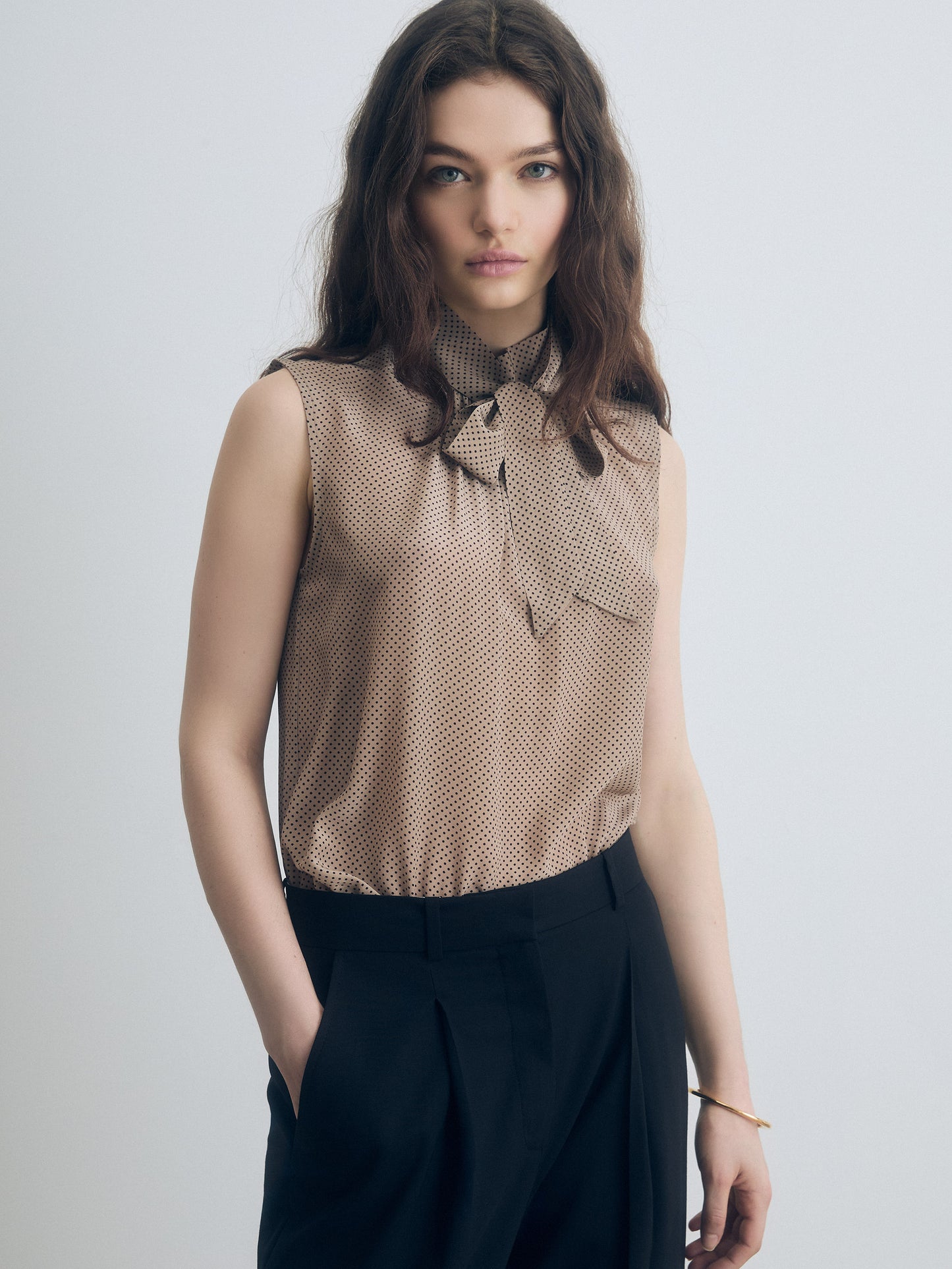 Sleeveless beige blouse with a subtle black micro-dot pattern, featuring a tie-neck detail and a single chest pocket, paired with high-waisted black pleated trousers.