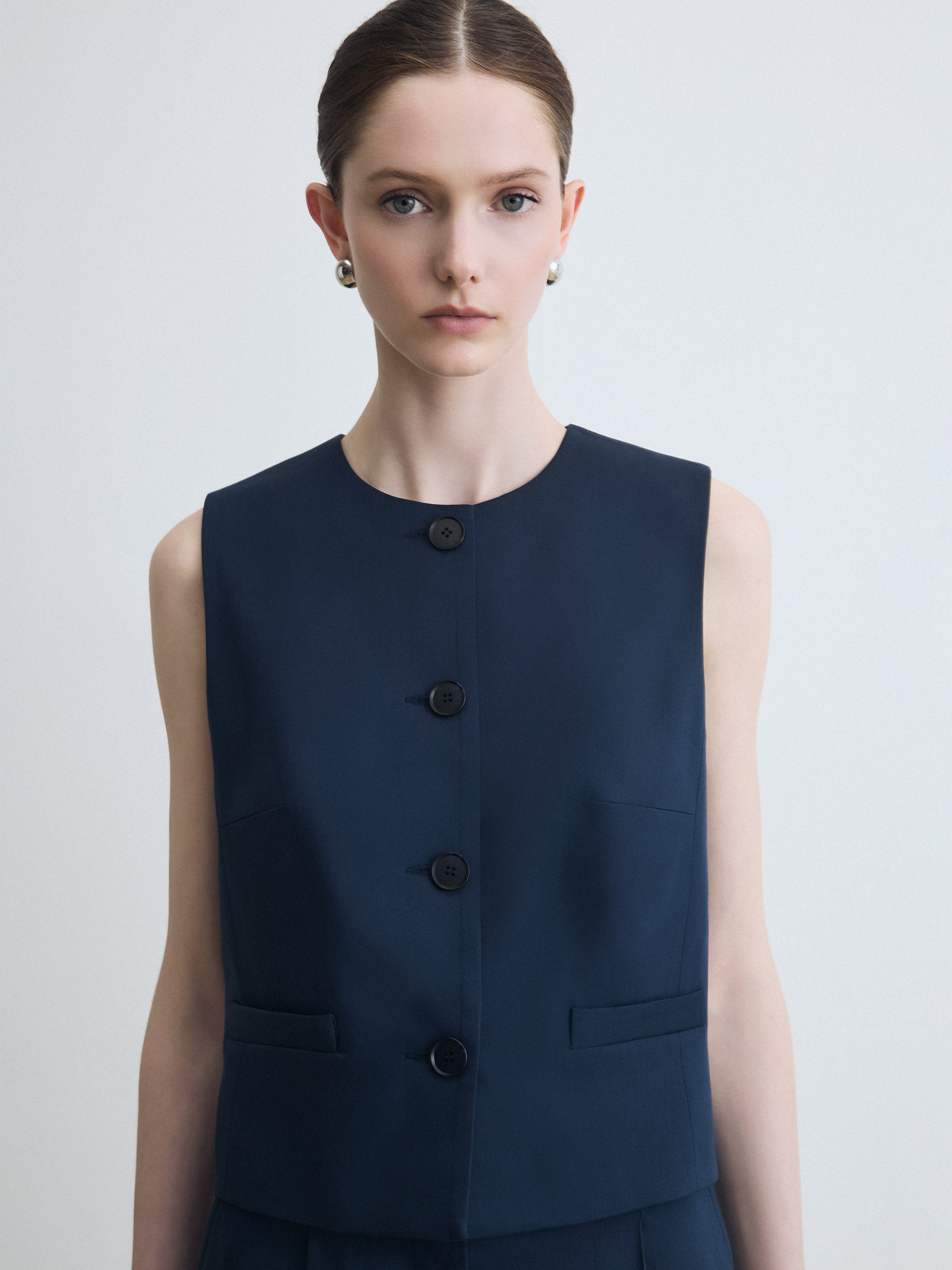 Woman wearing a navy blue sleeveless vest with buttons on a light gray background