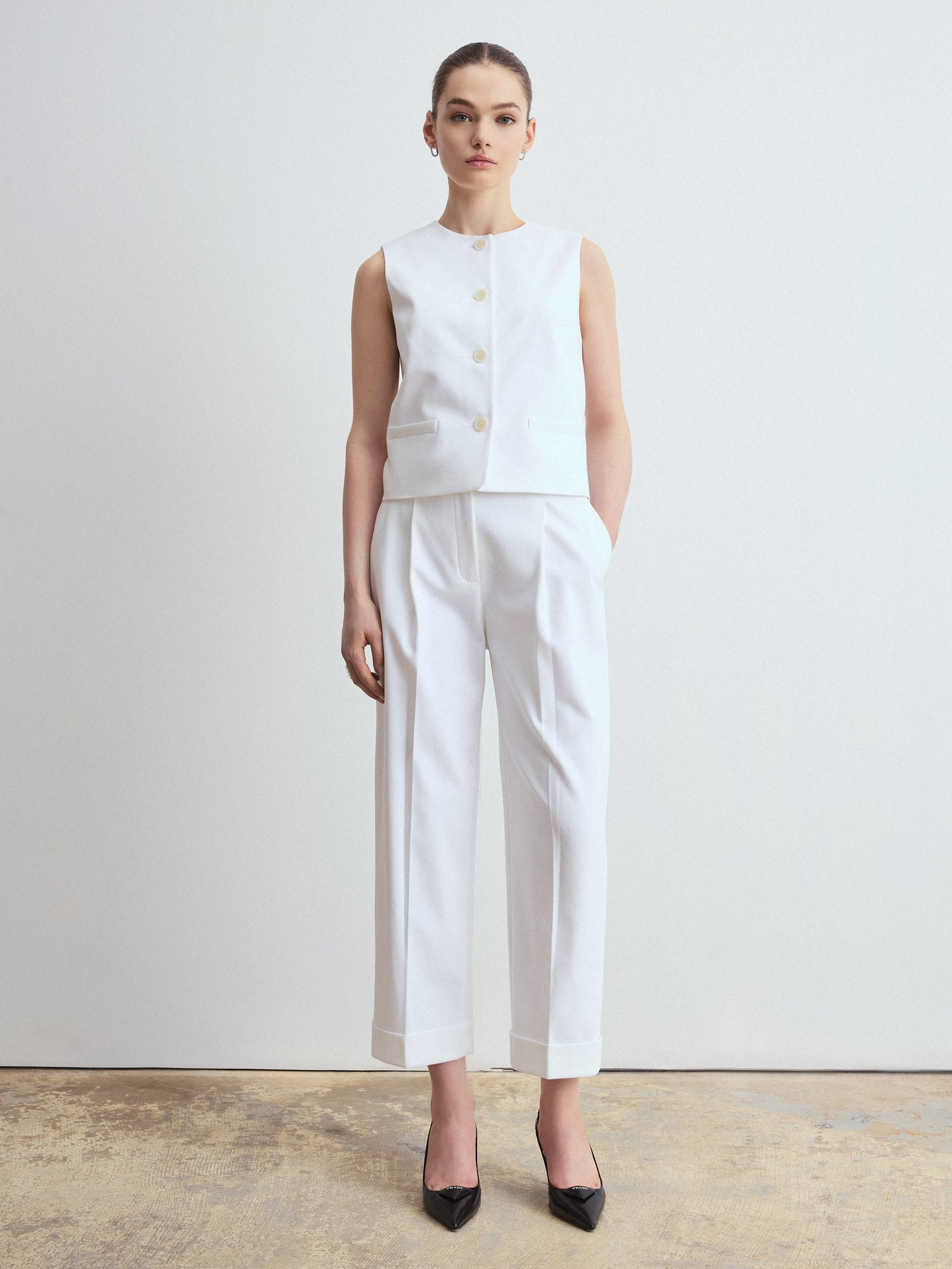 Person wearing a minimalist white sleeveless buttoned vest paired with matching tailored white trousers and black pointed-toe heels, standing against a plain light background.