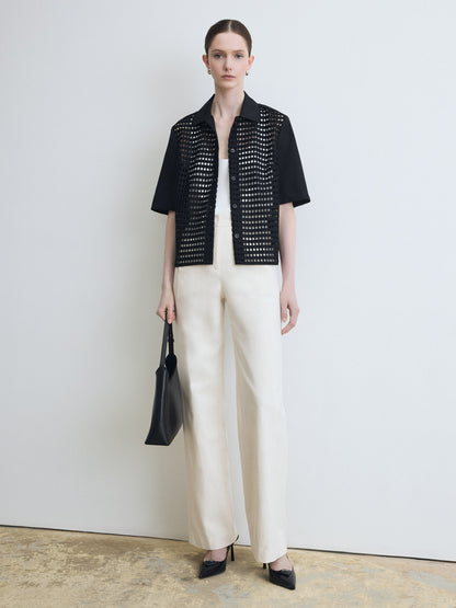 Woman wearing a black open-knit top and white pants, holding a black handbag against a plain background.