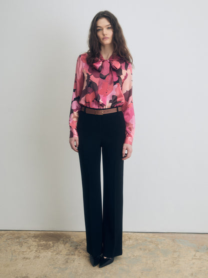 A person wearing a long-sleeve blouse with a bold floral print in shades of red, pink, and burgundy, featuring a bow tie at the neckline. The blouse is tucked into high-waisted black tailored trousers, paired with a slim tan belt and pointed black shoes. The background is a plain light wall with a neutral-toned floor
