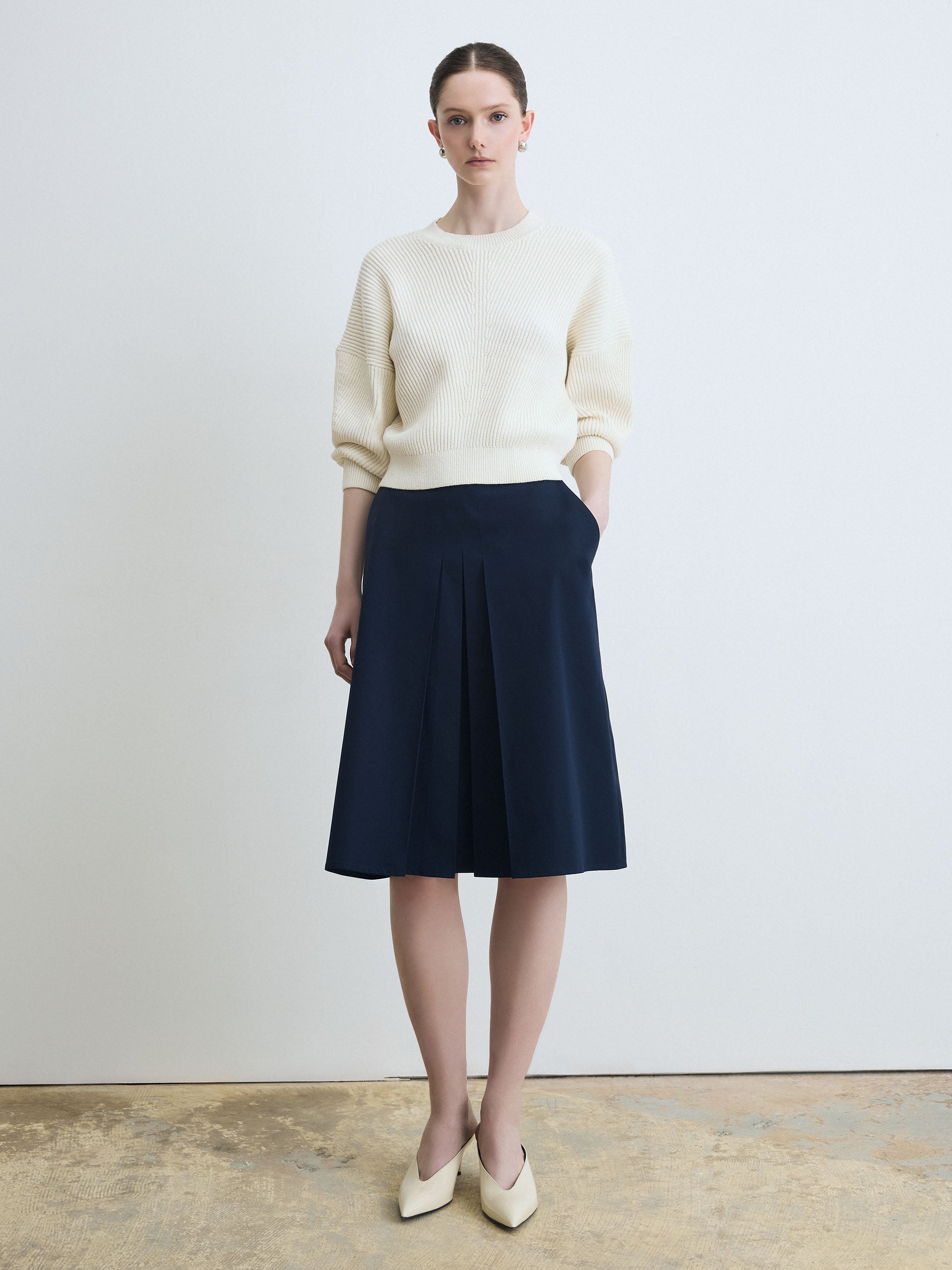 Woman wearing a cream sweater and navy skirt against a plain background