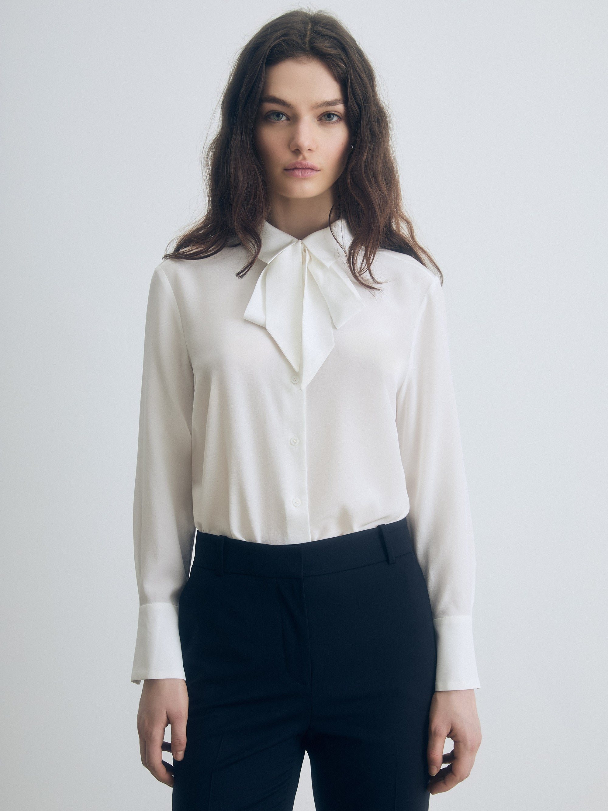 Woman wearing a white blouse with a bow tie and dark pants on a light gray background