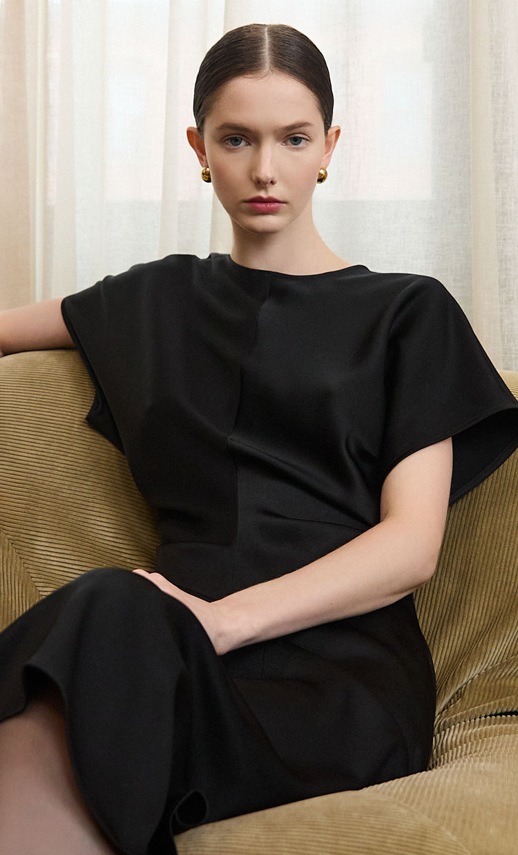 Mobile homepage banner featuring a person seated on a textured beige sofa, wearing an elegant black short-sleeve dress with a relaxed silhouette, in front of sheer cream curtains.