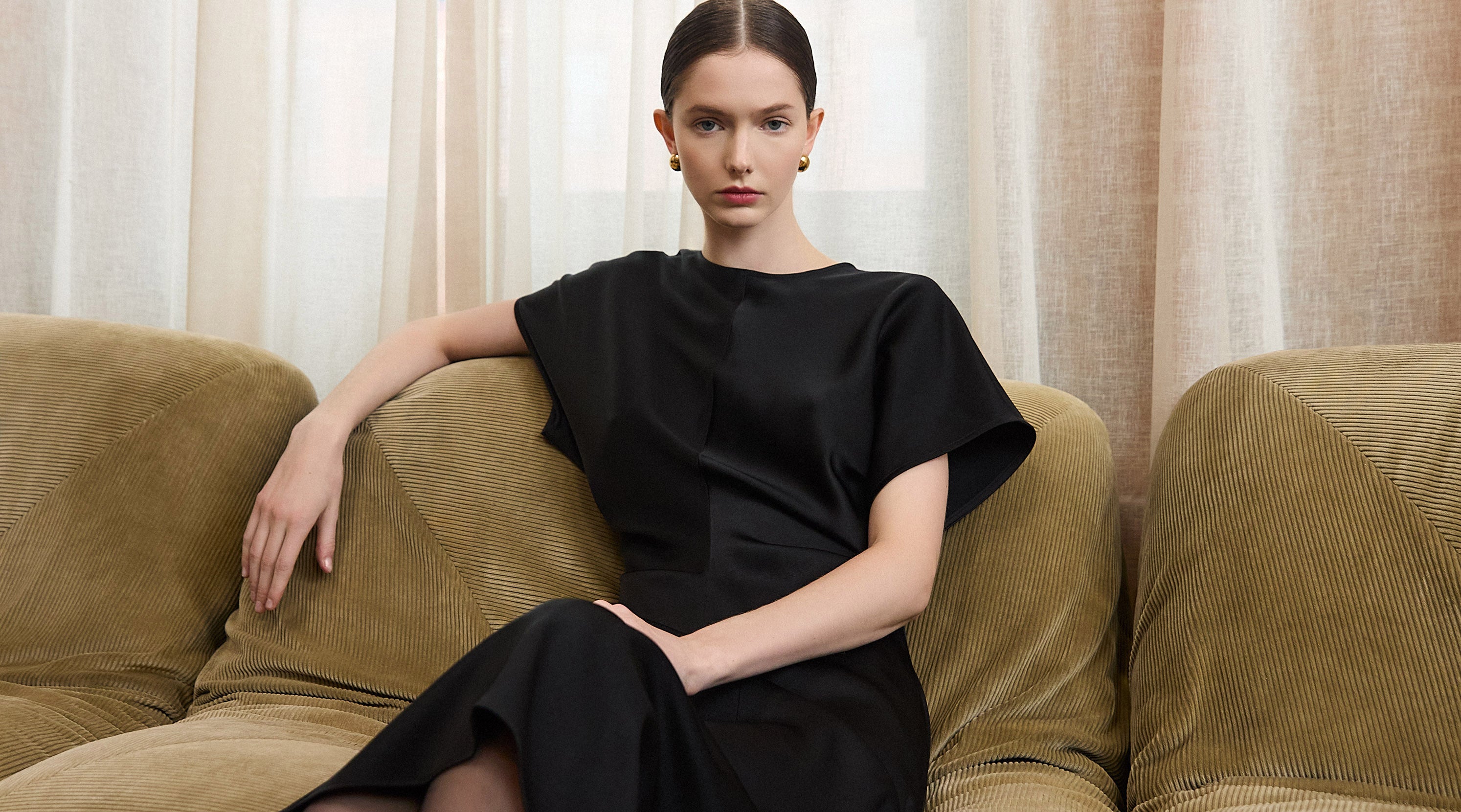 Judith & Charles homepage banner featuring a person seated on a textured beige sofa, wearing an elegant black short-sleeve dress with a fluid silhouette, in front of sheer neutral curtains.