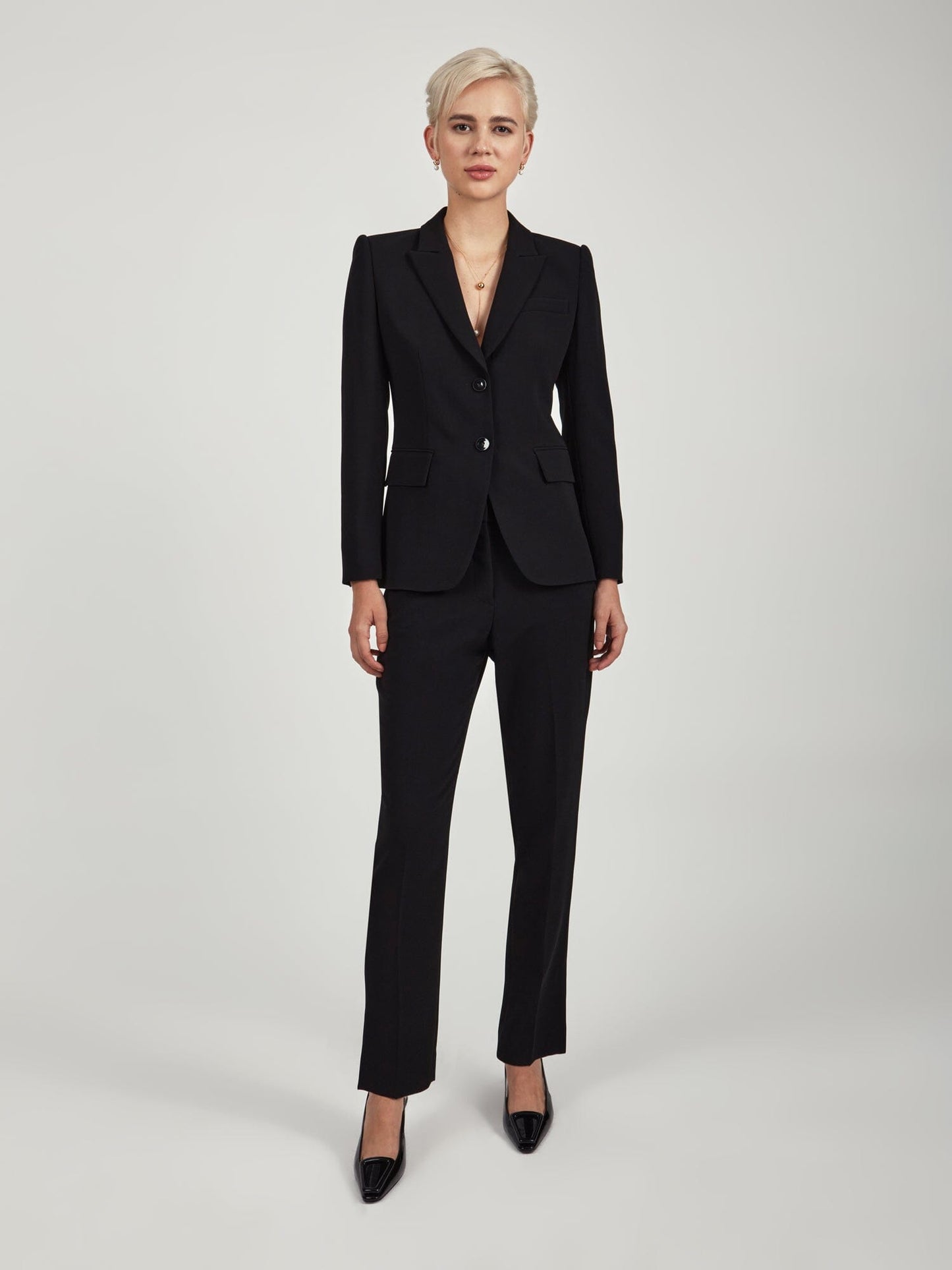 Model wearing slim ankle‑length triacetate trousers in black  tone, featuring a mid‑rise clean waist, slash pockets, back welt pockets, and a tailored slim leg. Styled with a matching blue blazer and black pointed‑toe heels against a minimal light grey background