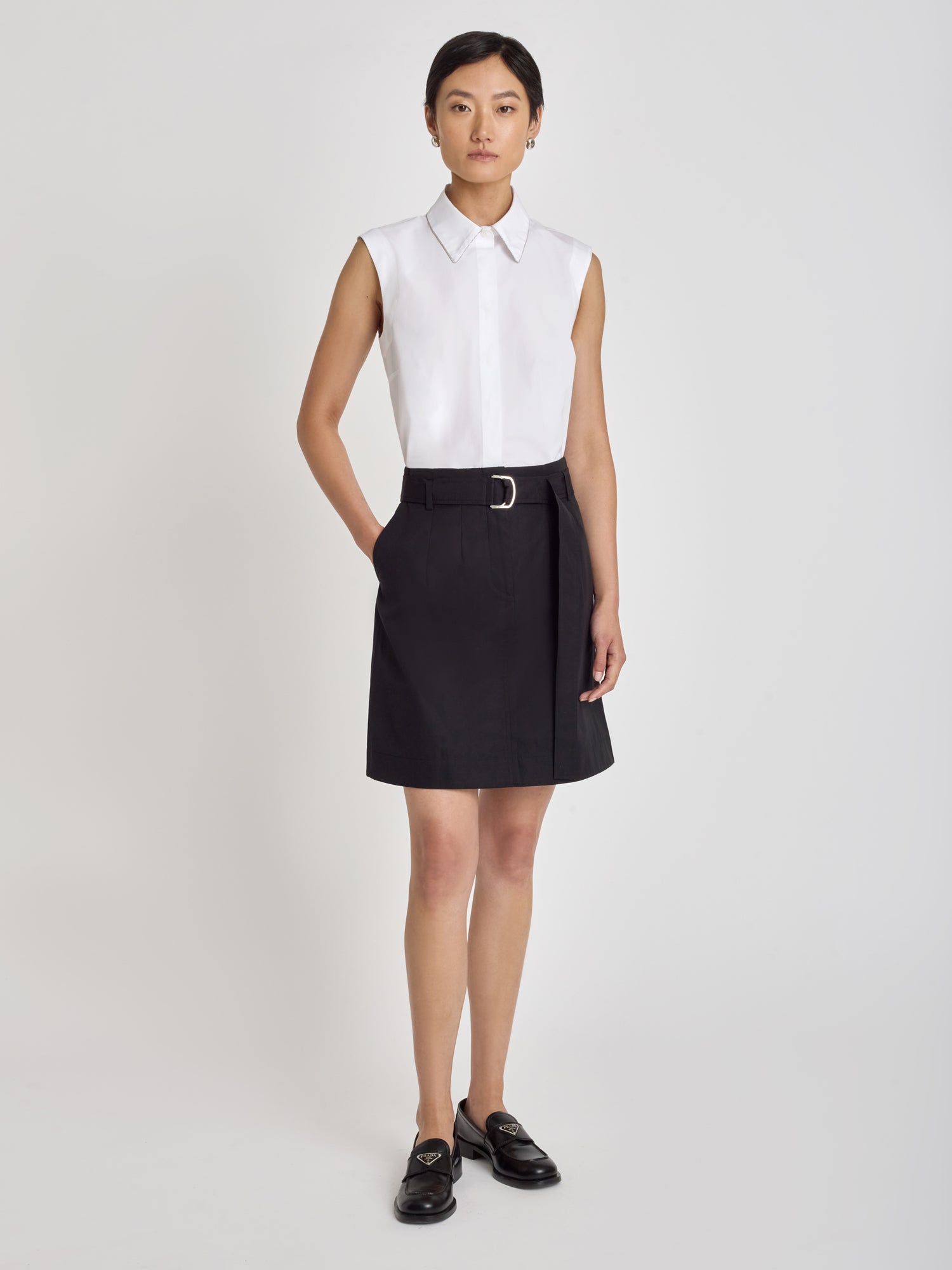 Amara structured trouser skirt in black with front zipper, slash pockets, and self belt with metal buckle, styled with a sleeveless white collared shirt and black loafers