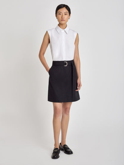 Amara structured trouser skirt in black with front zipper, slash pockets, and self belt with metal buckle, styled with a sleeveless white collared shirt and black loafers
