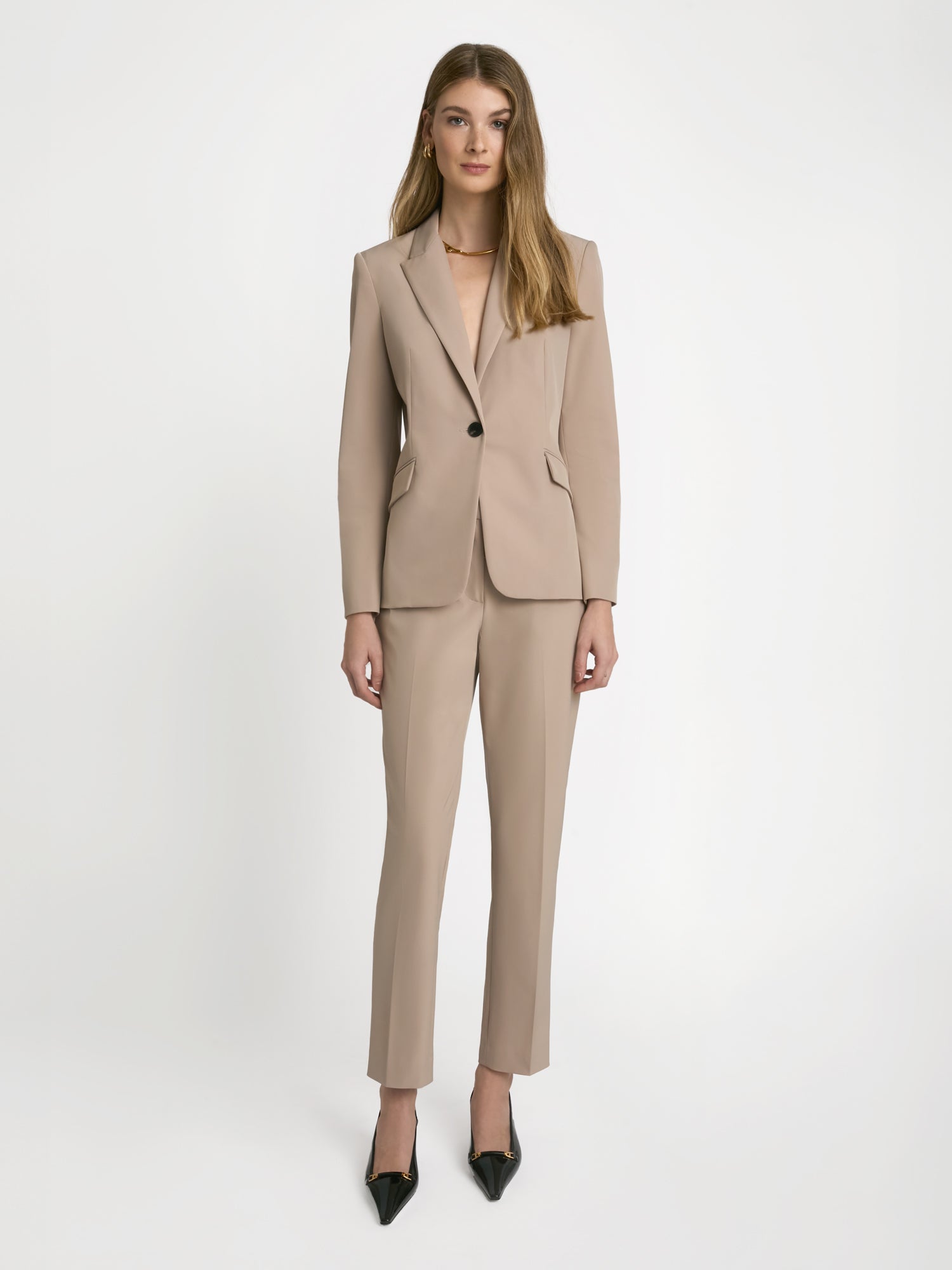 A model wearing ankle length slim pants with mid-rise waist, featuring slash pockets, back welt pockets, hook and eye closure with zip, and belt loops in a beige color.