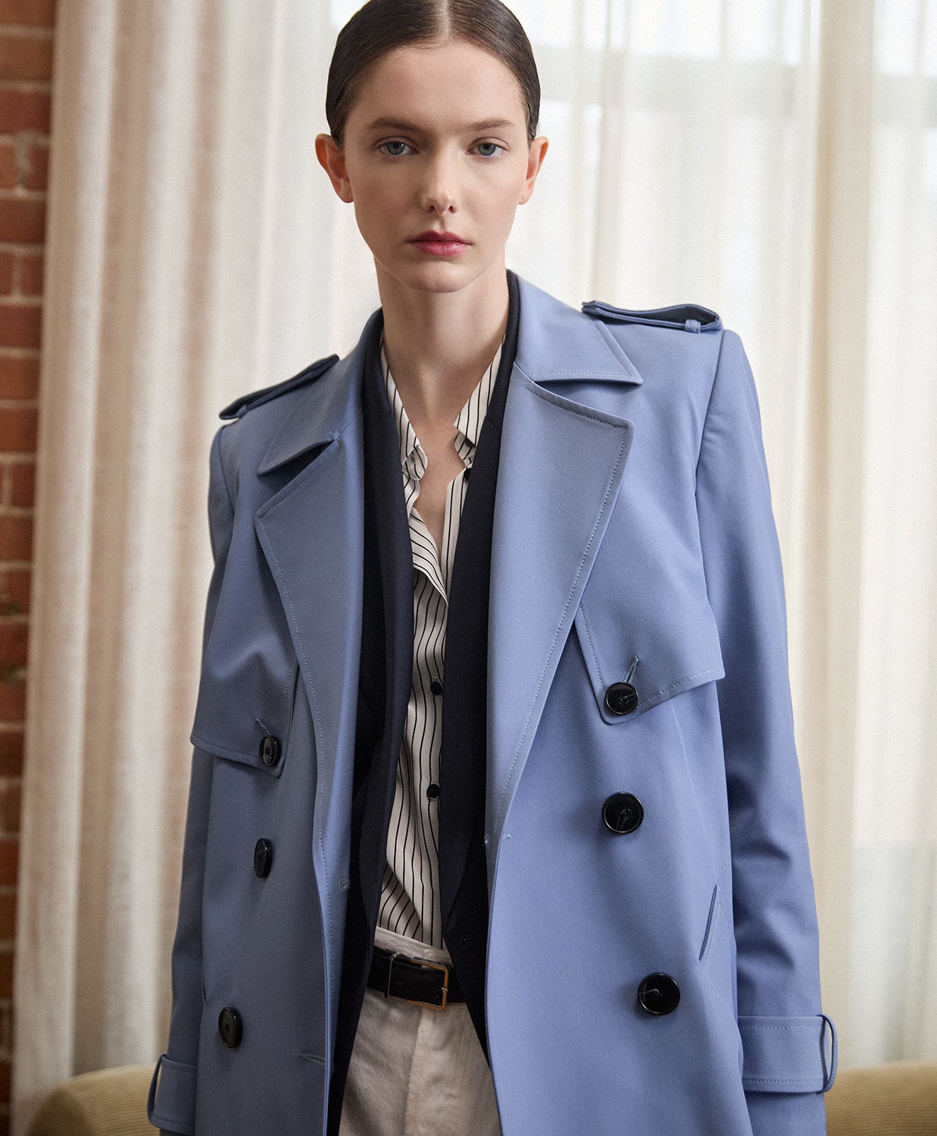 Light blue double-breasted trench coat layered over a tailored navy blazer and a white shirt with black pinstripes, styled with light trousers.